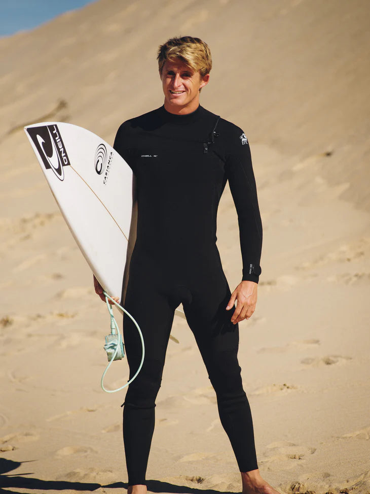 Man in a black Oneill epic wetsuit holding a white surfboard on sandy terrain